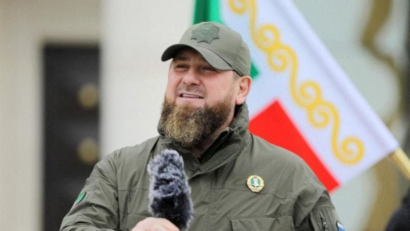 Ramzan Kadyrov in military uniform and a cap, speaking into a microphone with a flag in the background.