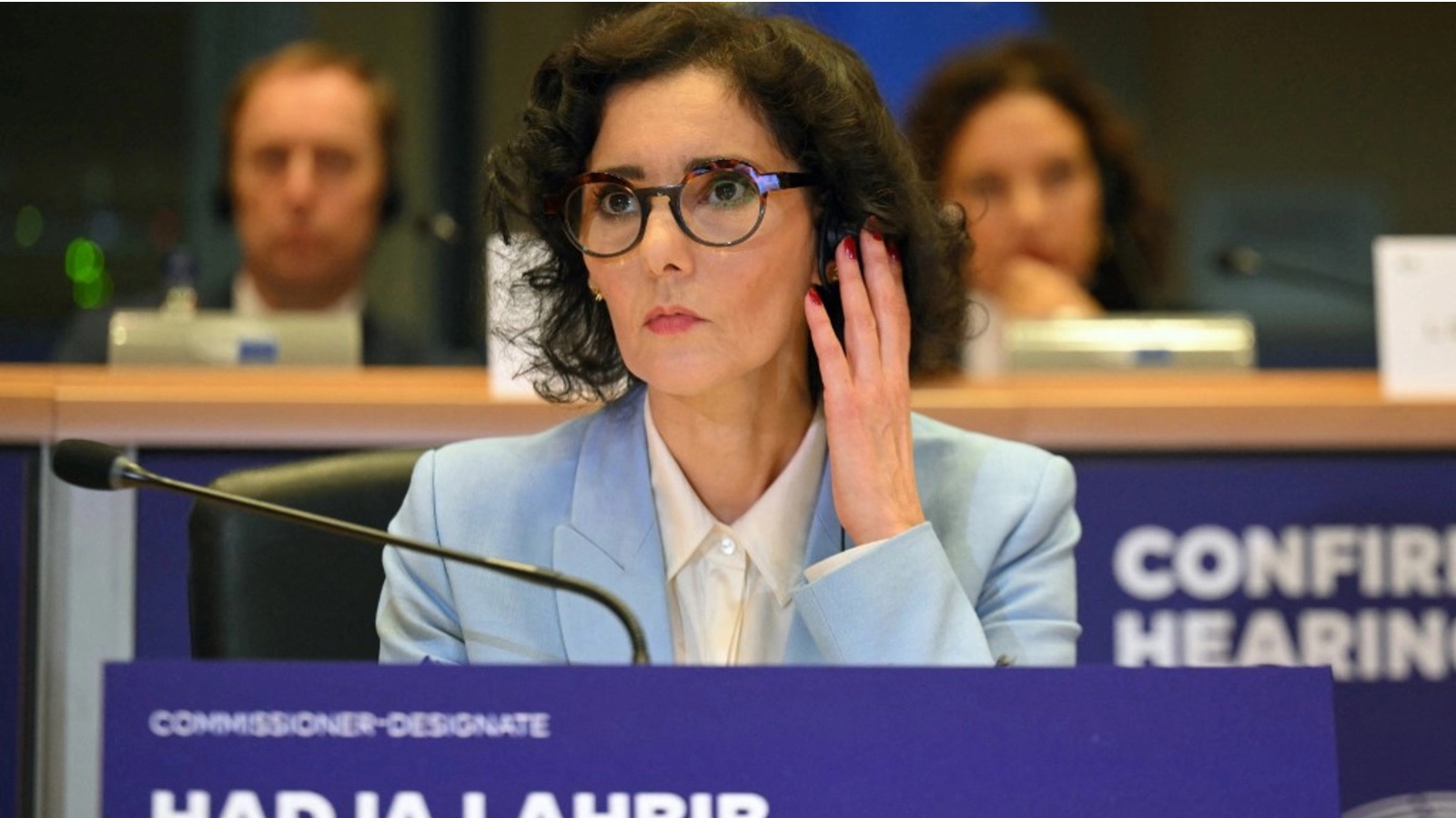 Hadja Lahbib wearing glasses and a light blue suit, listening through an earpiece during a hearing at the European Parliament.