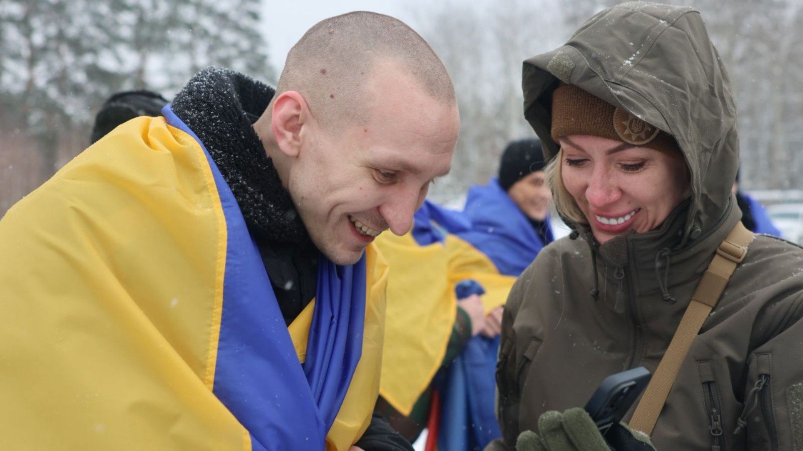 A smiling released soldier wrapped in the Ukrainian flag looks at a phone with a woman in military uniform against a snowy background.