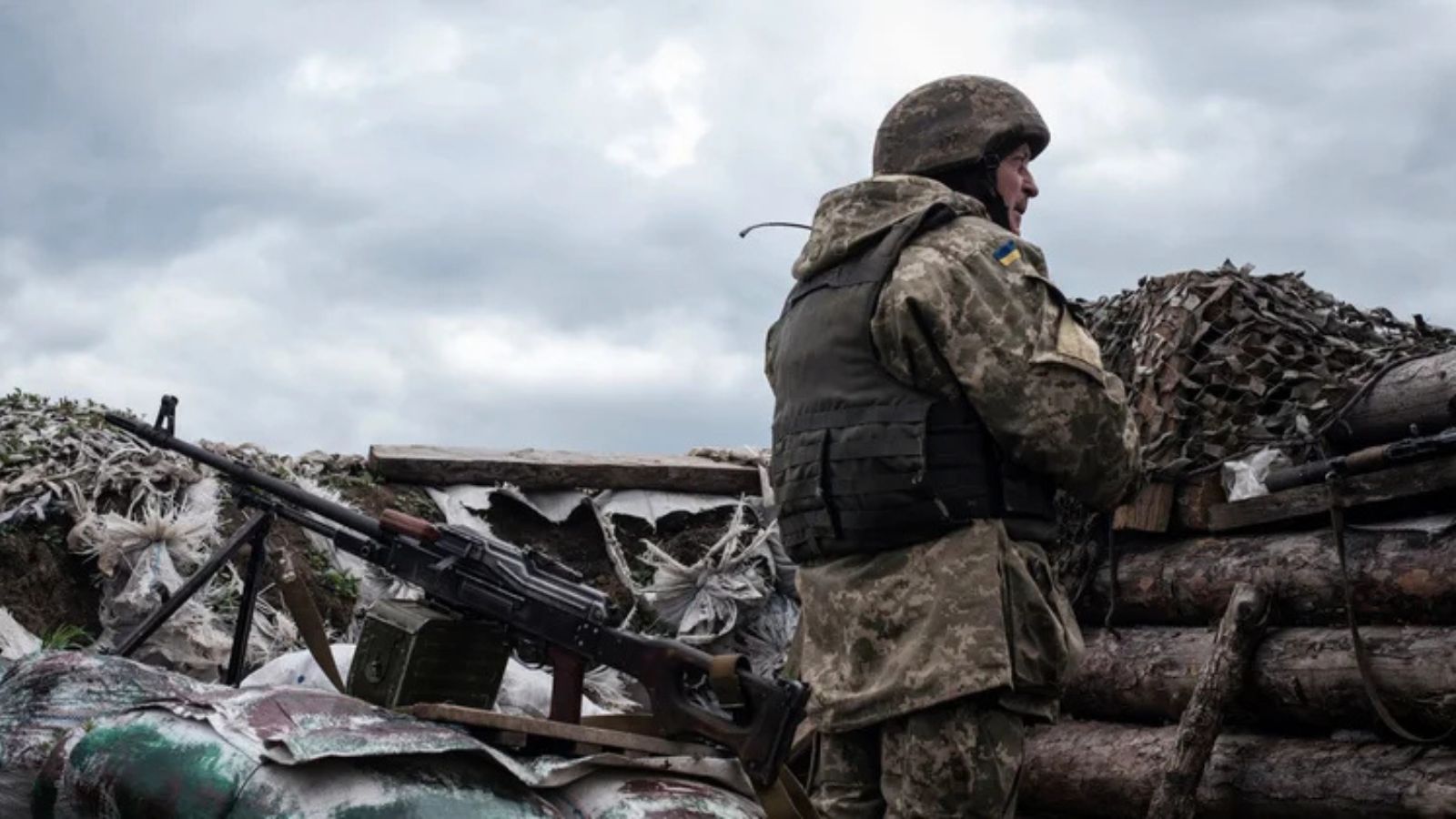 A Ukrainian defender in camouflage and a bulletproof vest stands at a fighting position in a reinforced trench next to a machine gun.