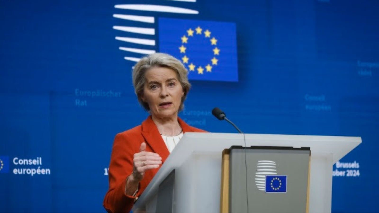Ursula von der Leyen speaking at a podium with a blue European Union flag background.