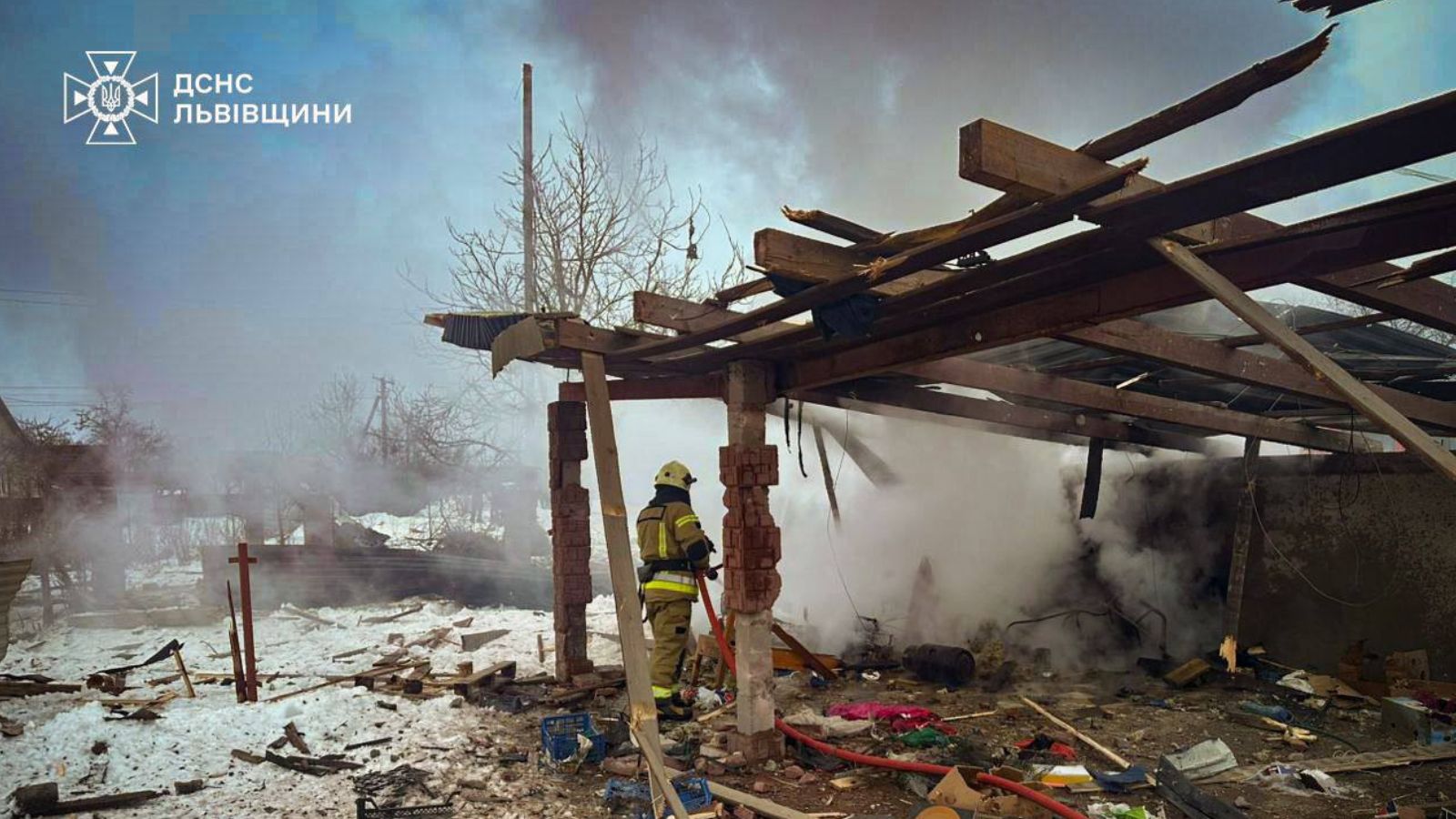 A rescuer stands among the destroyed wooden and brick structures of a civilian building, with thick smoke rising after the shelling