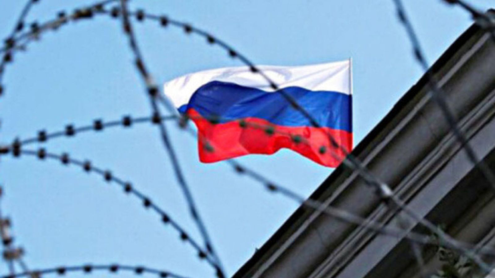 The Russian tricolor flag waving against a blue sky, viewed through barbed wire and a metal fence