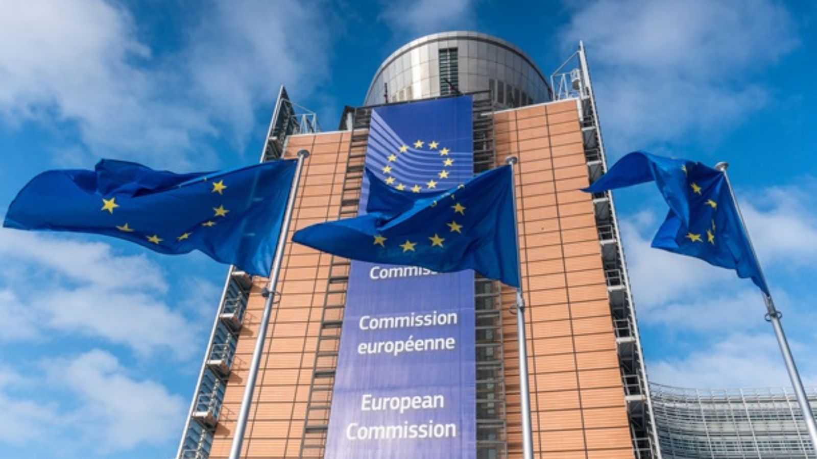 Low-angle shot of the European Commission building in Brussels with EU flags waving against a blue sky.