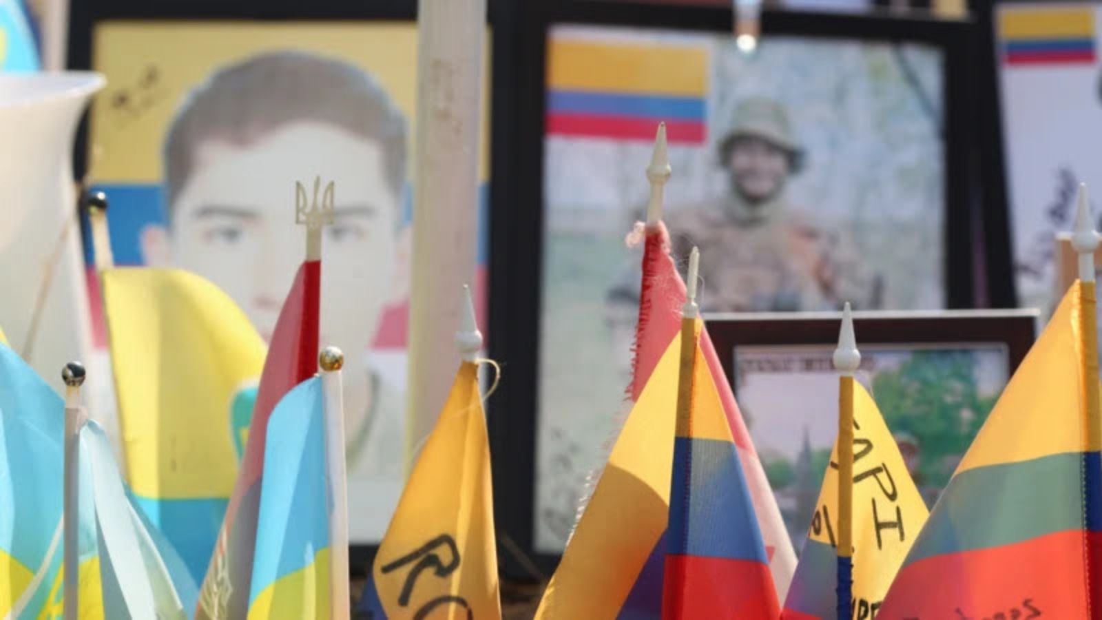 A close-up of a memorial site at Maidan Nezalezhnosti showing framed portraits of fallen soldiers and small Colombian and Ukrainian flags.