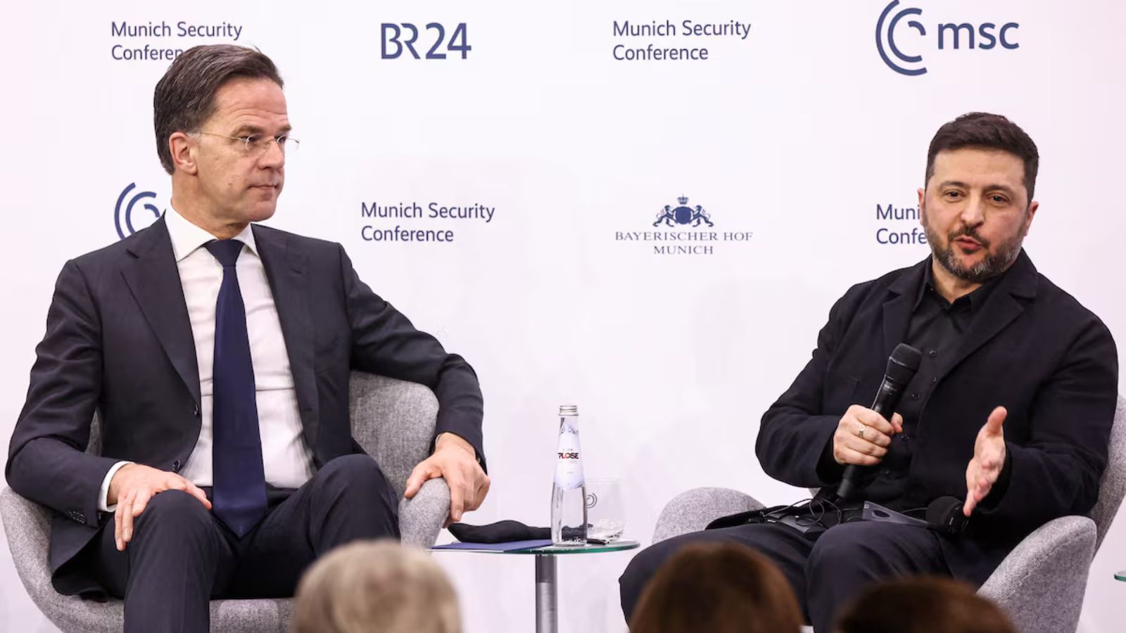 Mark Rutte and Volodymyr Zelenskyy sitting in armchairs during a panel discussion at the Munich Security Conference. Zelenskyy is holding a microphone and gesturing while speaking.