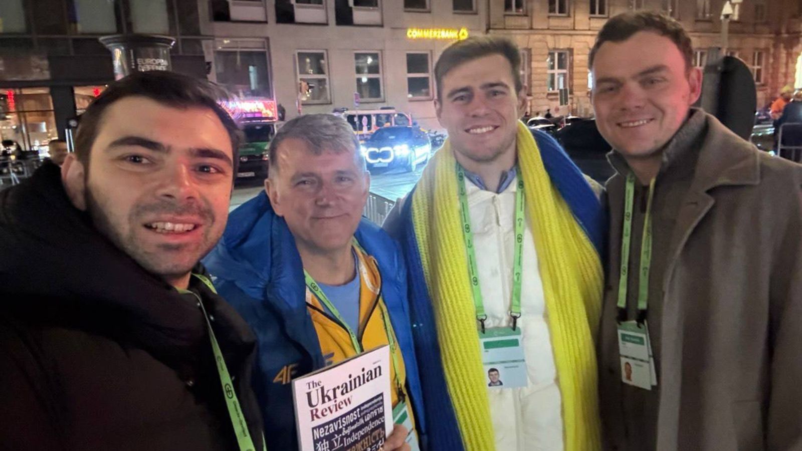 A selfie of four men on a Munich street at night; one is holding a copy of The Ukrainian Review magazine.