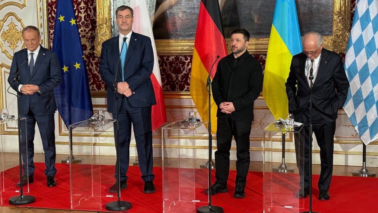 Four politicians standing at microphones on a red carpet in front of the flags of the EU, Germany, Ukraine, and Bavaria.