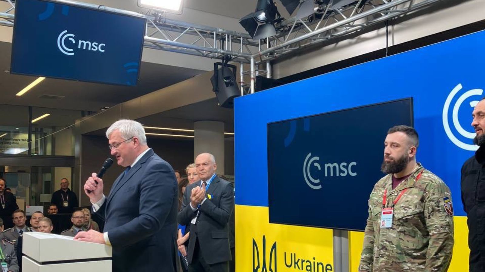 Andrii Sybiha speaking at a lectern with a microphone, with military personnel and officials standing nearby against a blue and yellow banner.