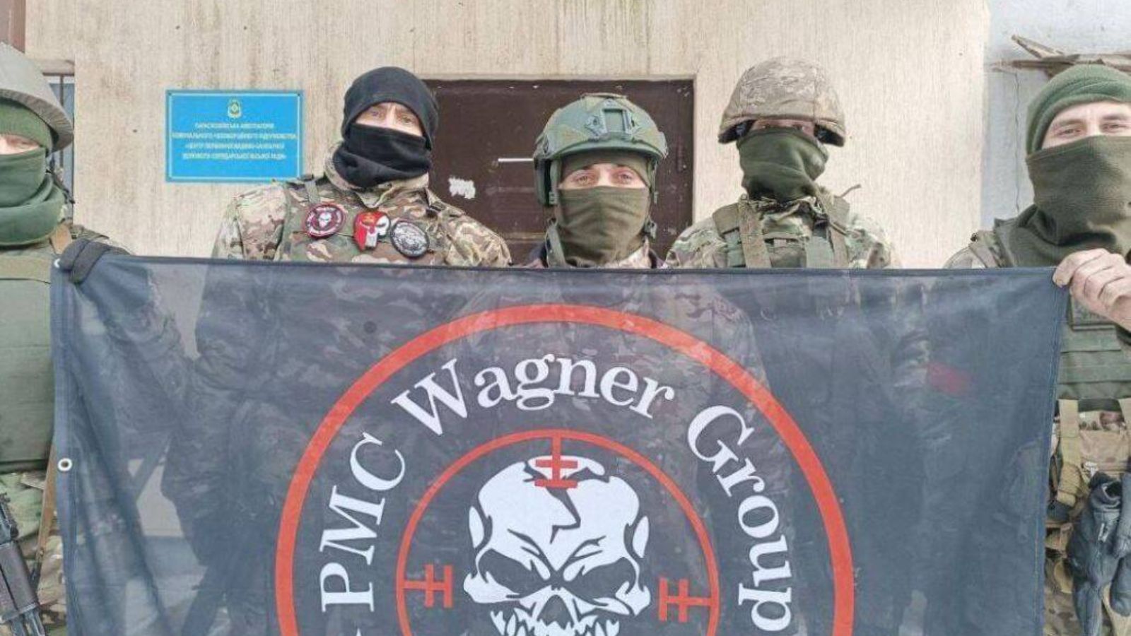 Armed men in camouflage wearing Wagner Group patches standing in front of military vehicles