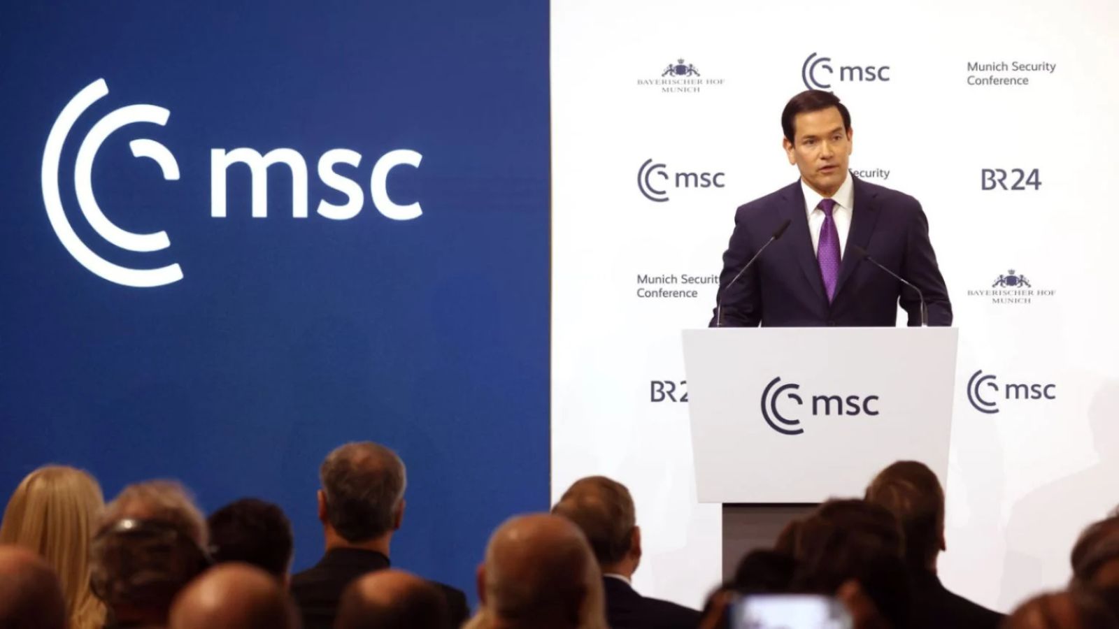 Marco Rubio speaking at a white podium in front of an audience against a blue and white MSC banner.