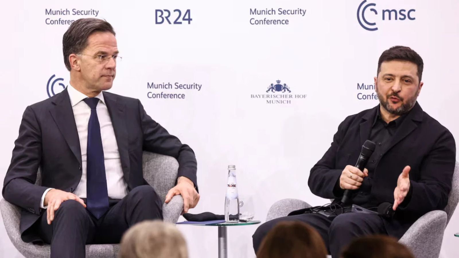 Mark Rutte and Volodymyr Zelenskyy sitting in armchairs during a panel discussion; Zelenskyy is holding a microphone and gesturing.