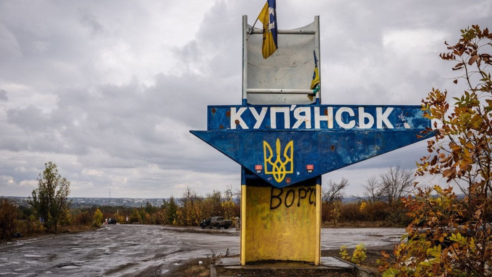 A road sign with the inscription "Kupiansk" in blue and yellow colors, featuring the Ukrainian coat of arms and flag against a background of a road and cloudy sky.