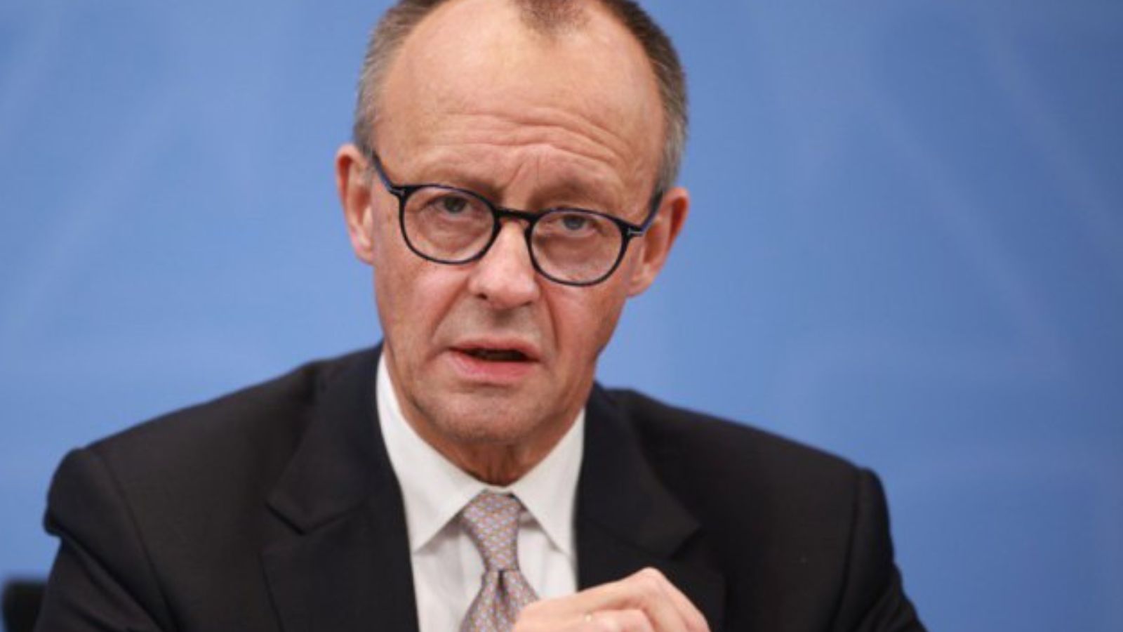 Close-up portrait of German Chancellor Friedrich Merz wearing glasses and a business suit against a blue background