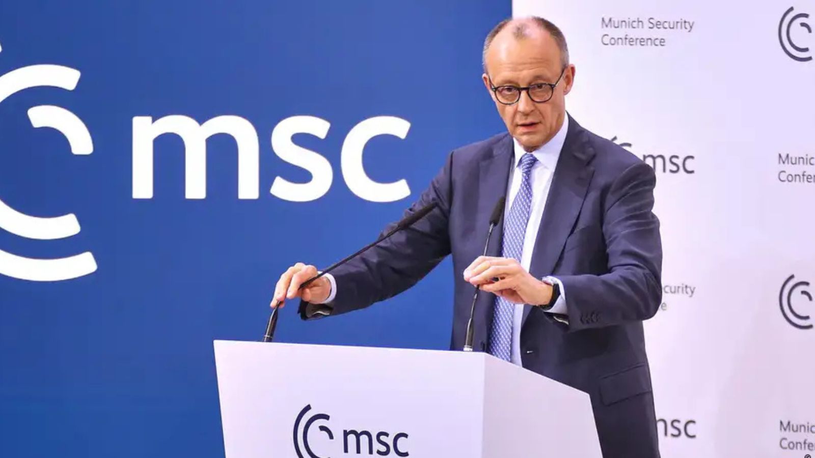 Friedrich Merz in glasses and a blue suit speaking at a podium, holding the edges of the microphones