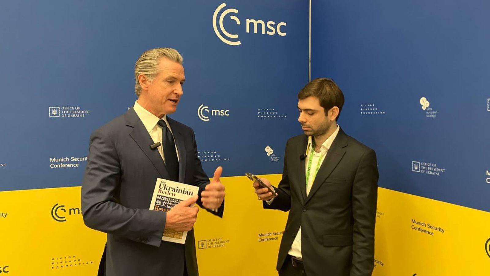 Gavin Newsom holding The Ukrainian Review magazine and talking to an editorial representative in front of an MSC banner