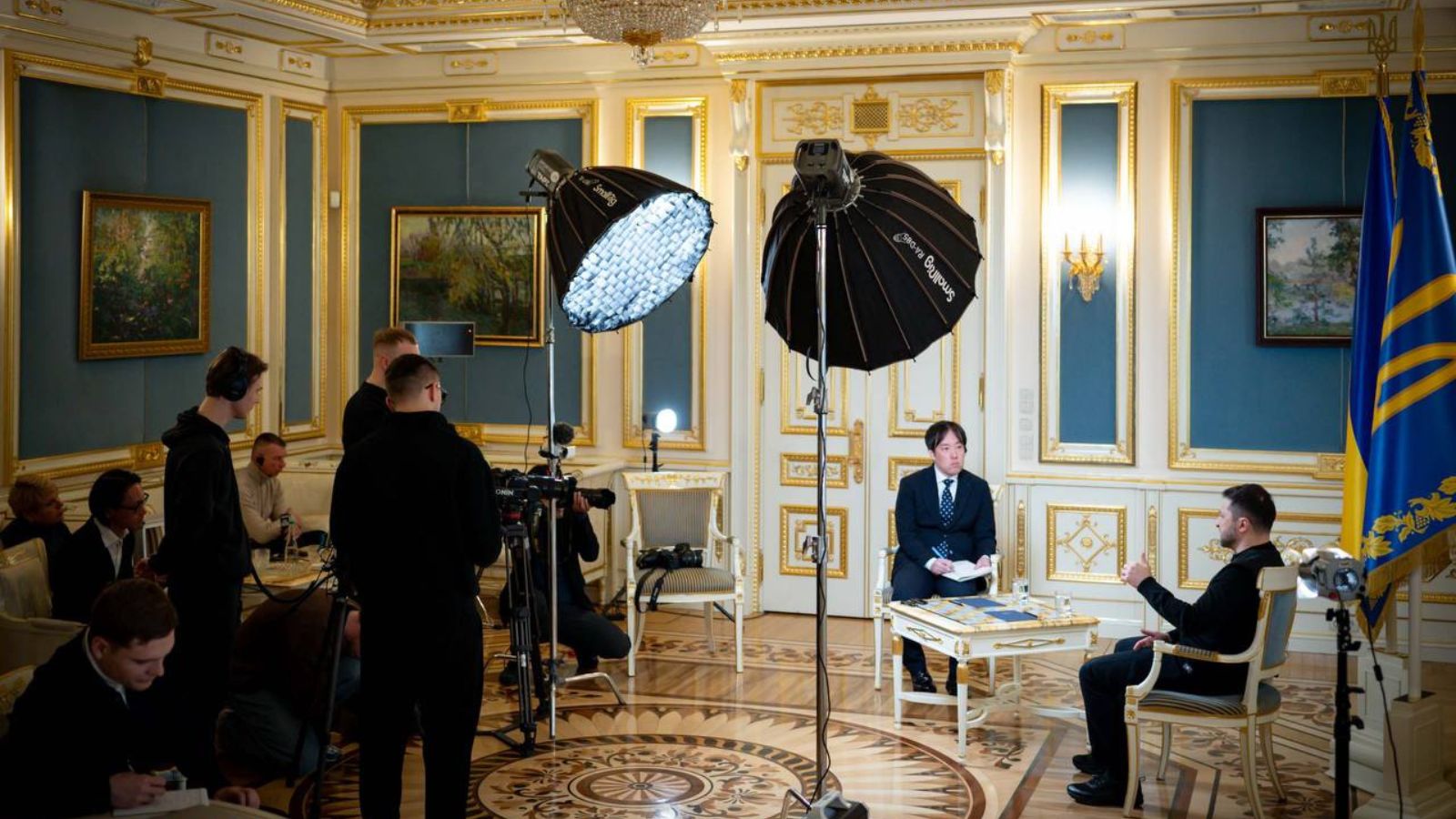 Volodymyr Zelenskyy giving an interview to a Japanese journalist in an official hall, surrounded by a film crew and lighting equipment.