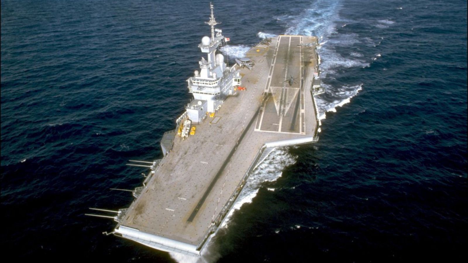High-angle view of the flight deck of the Charles de Gaulle aircraft carrier cruising through the ocean, leaving a white wake.