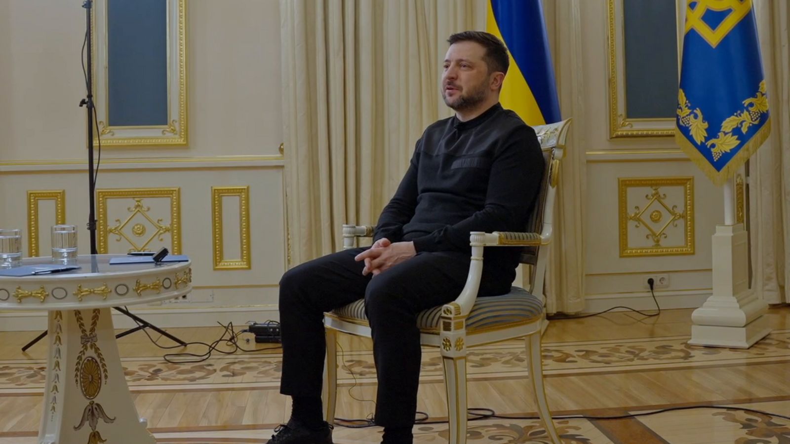 Volodymyr Zelenskyy, dressed in black, sits in a classic white chair against the backdrop of the Ukrainian flag during a conversation with journalists.