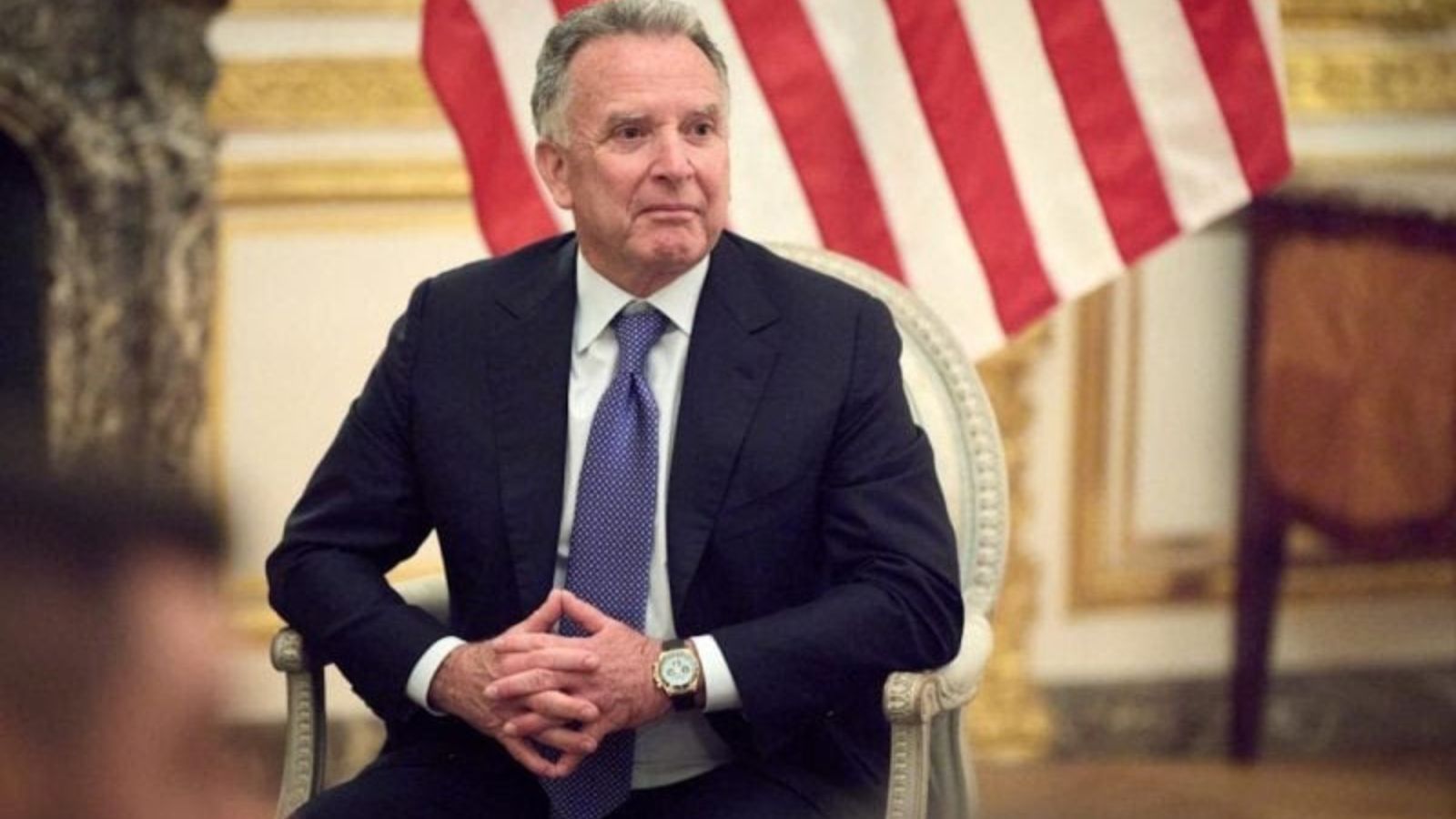 Steven Witkoff sitting in a chair during an official meeting with the American flag behind him.