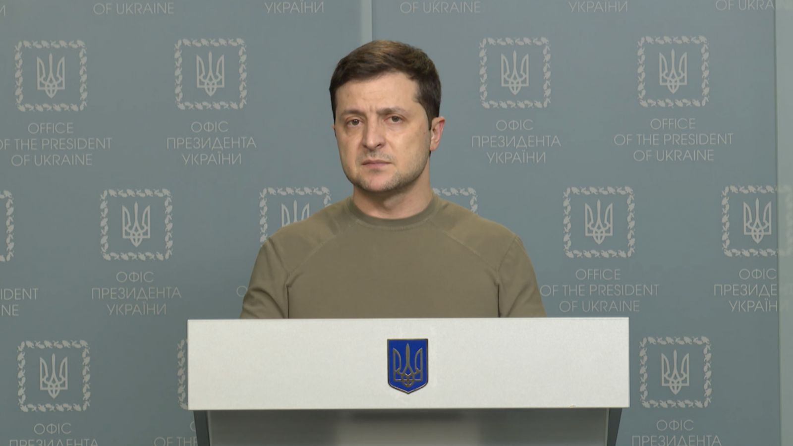 Volodymyr Zelenskyy in a khaki military t-shirt standing behind a lectern with the Ukrainian coat of arms against Office of the President backdrops.