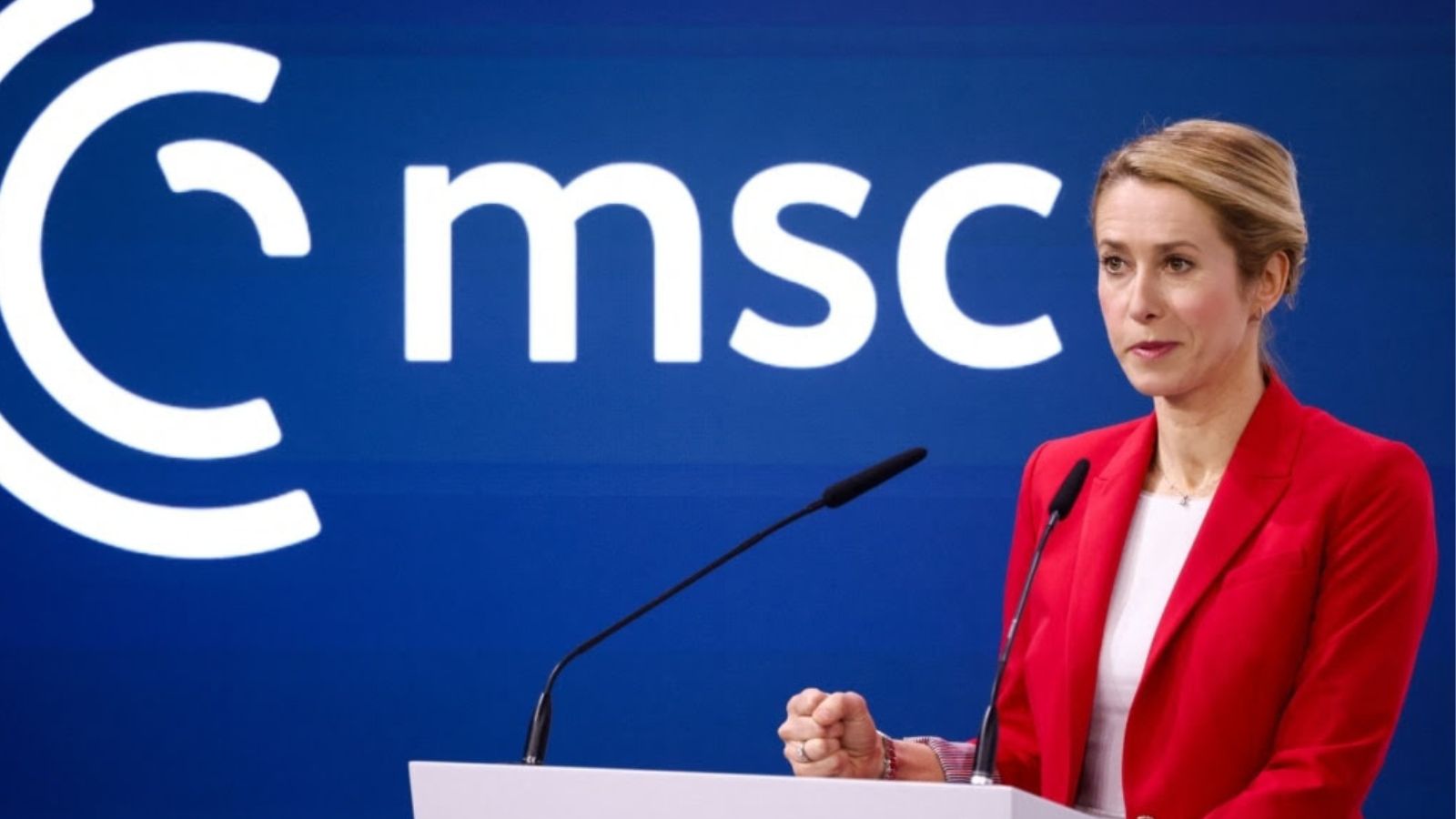 Kaja Kallas in a bright red blazer speaking at a podium in front of a blue banner with a large MSC logo.