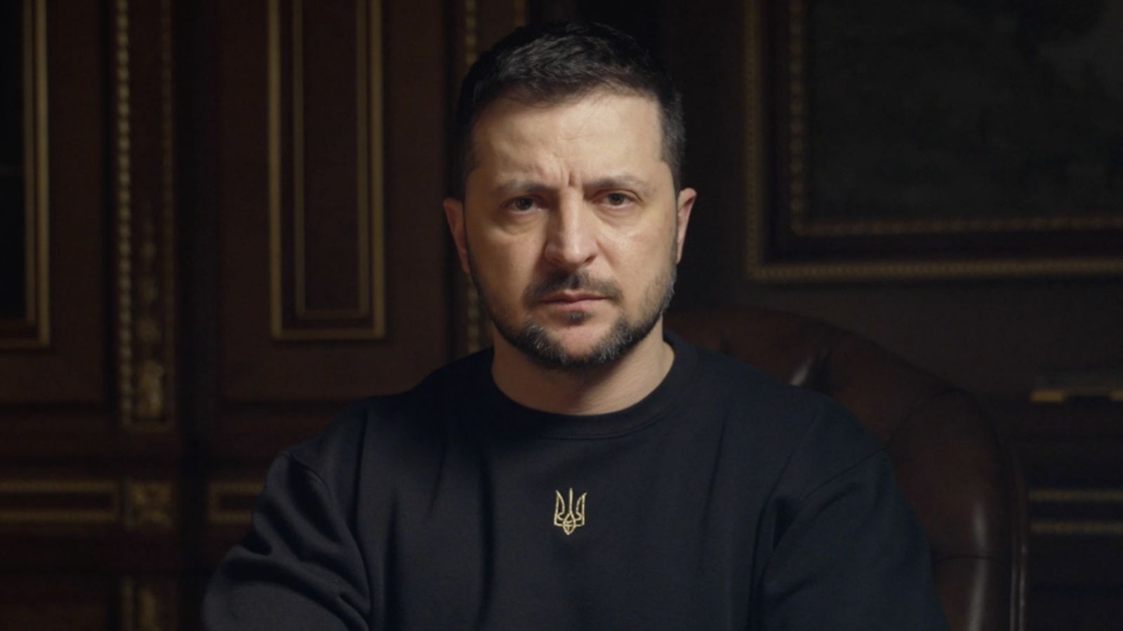 Volodymyr Zelenskyy in a black sweatshirt with a small trident, with a focused gaze against a dark office background.