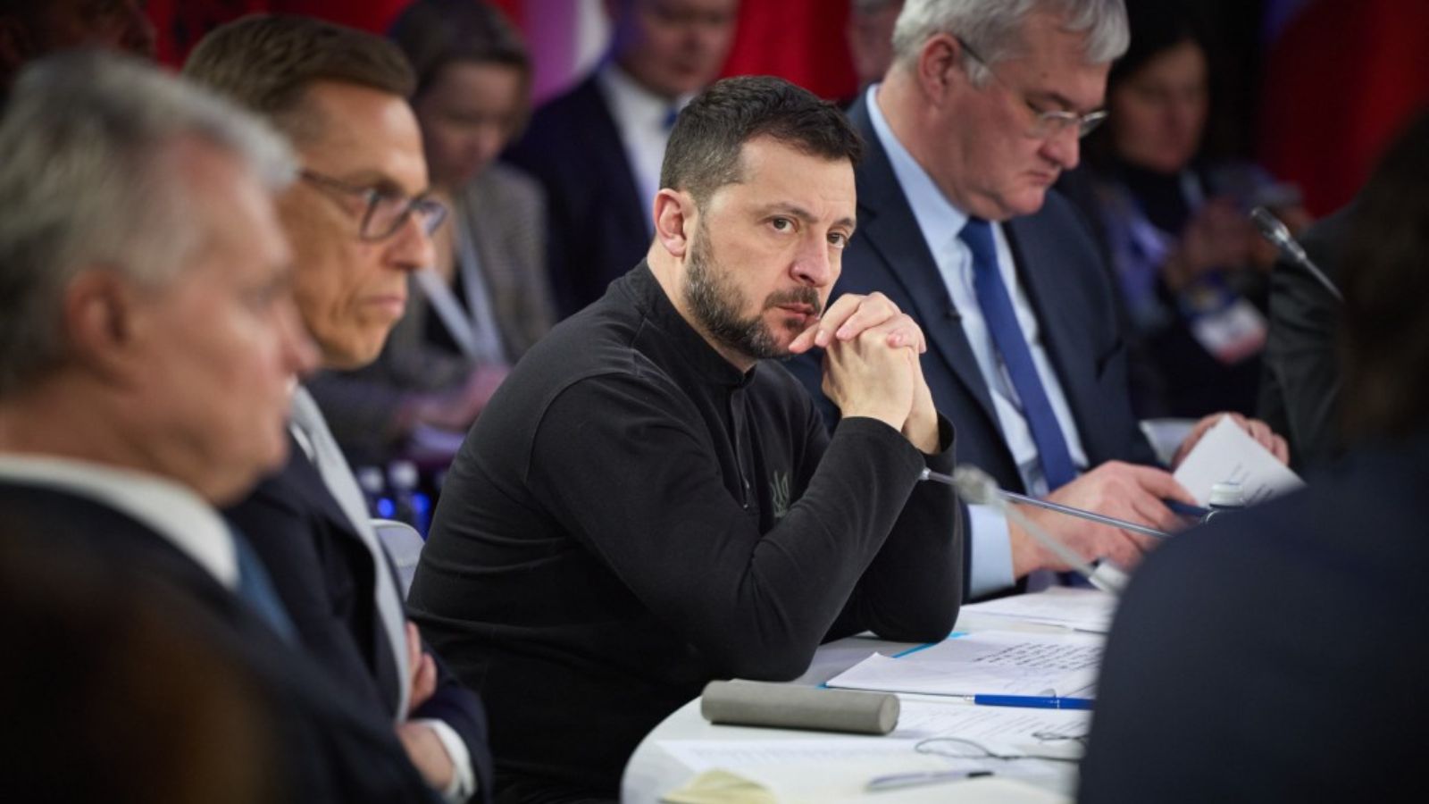 President Zelenskyy at the negotiating table with international partners, hands clasped, with a serious and determined expression.