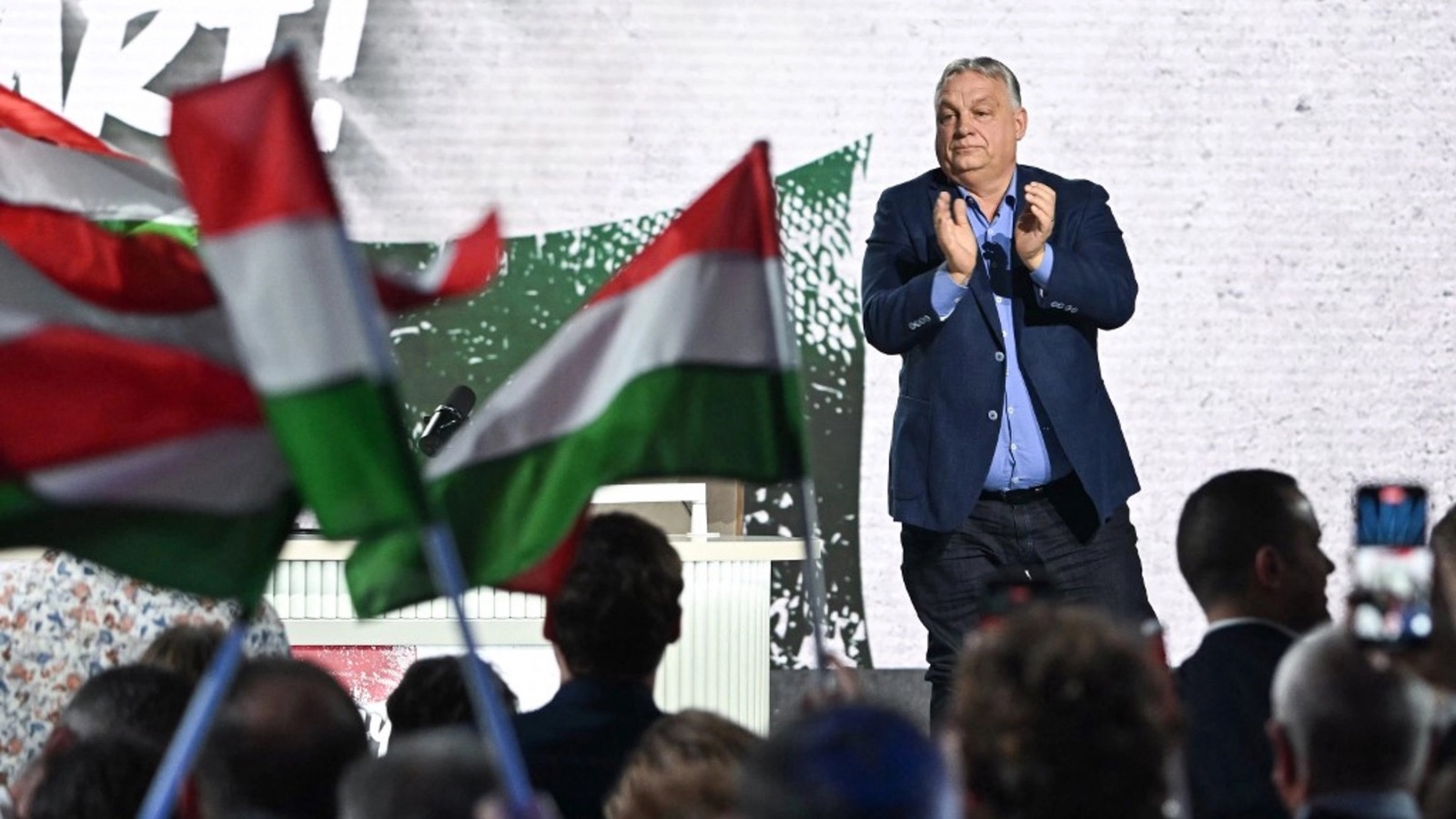 Віктор Орбан аплодує на сцені перед прихильниками на тлі угорських прапорів.