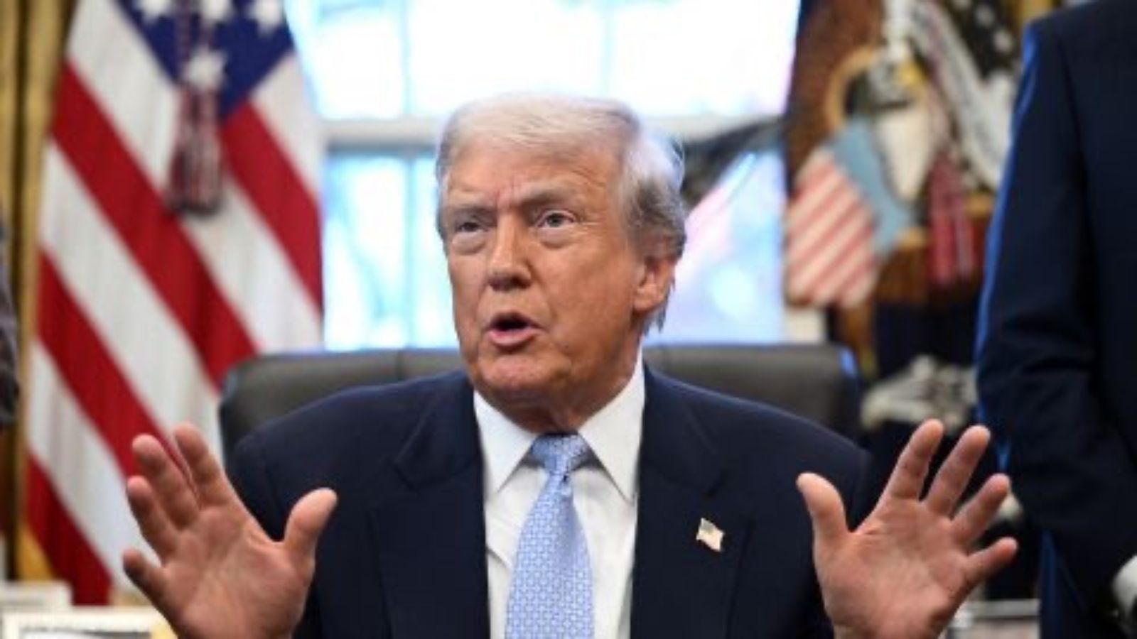 A photograph of Donald Trump sitting at a table and gesturing with his hands while speaking