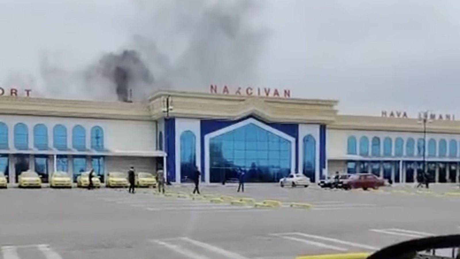 Screenshot of a video showing the Nakhchivan International Airport building with blue windows and gold Cyrillic lettering that says "NAXCIVAN." Thick black smoke is billowing from the roof of the building. Several yellow taxis and people are visible in the parking lot in front.