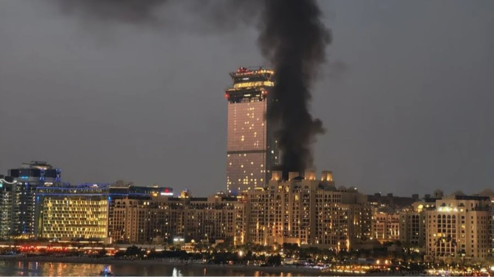View of Dubai at night with thick black smoke rising from a building near a skyscraper.