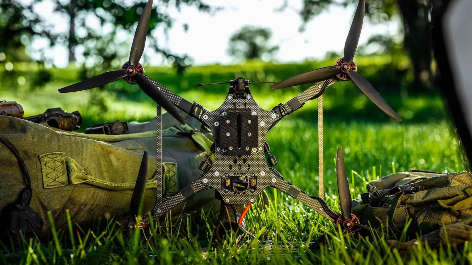 A modern carbon fiber quadcopter drone set against a background of green grass and military gear.