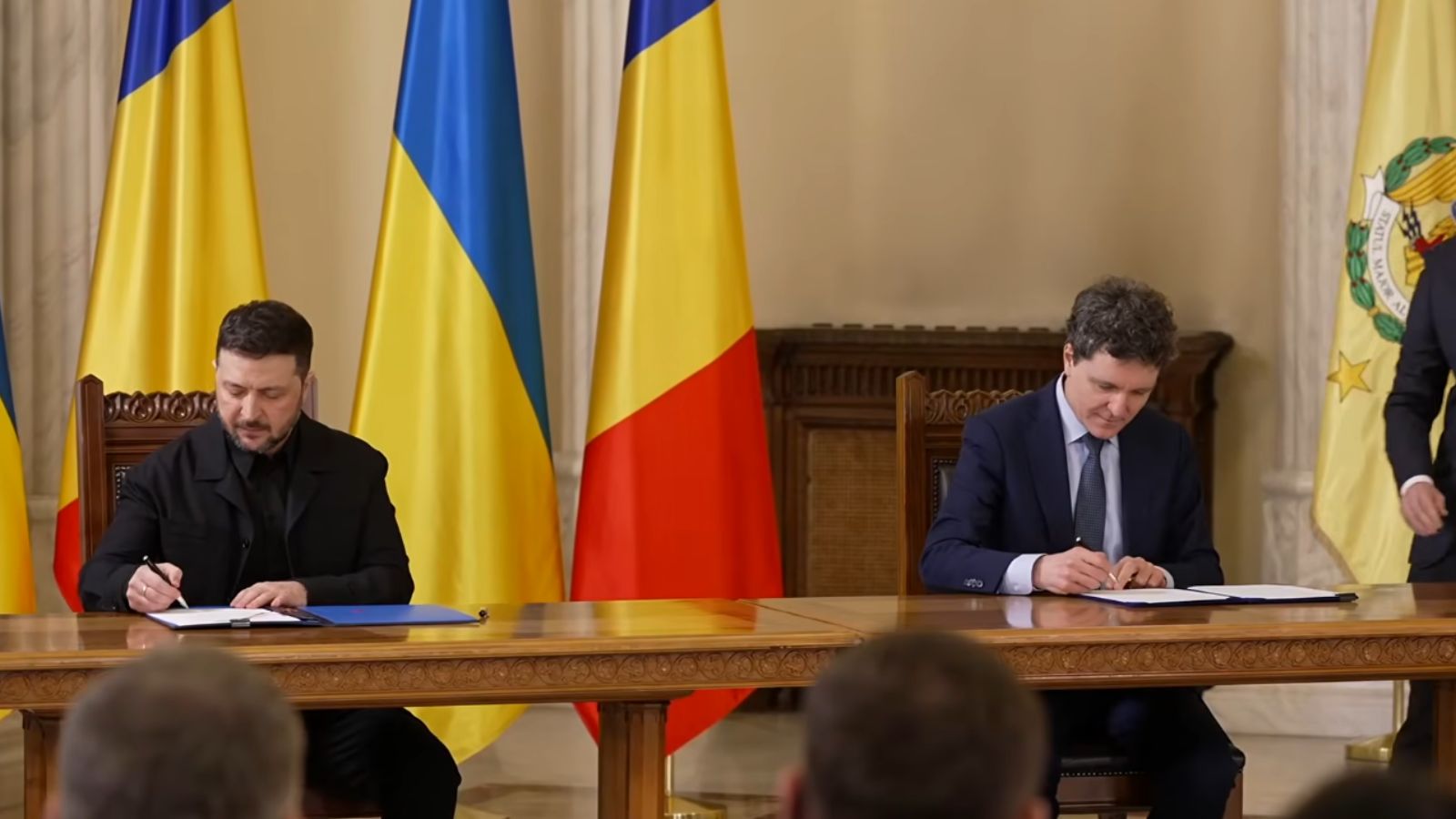 Президент України Володимир Зеленський та мер Бухареста Нікушор Дан потискують руки, стоячи. Нікушор Дан тримає підписану синю папку з гербом. Позаду них стоять прапори України та Румунії.