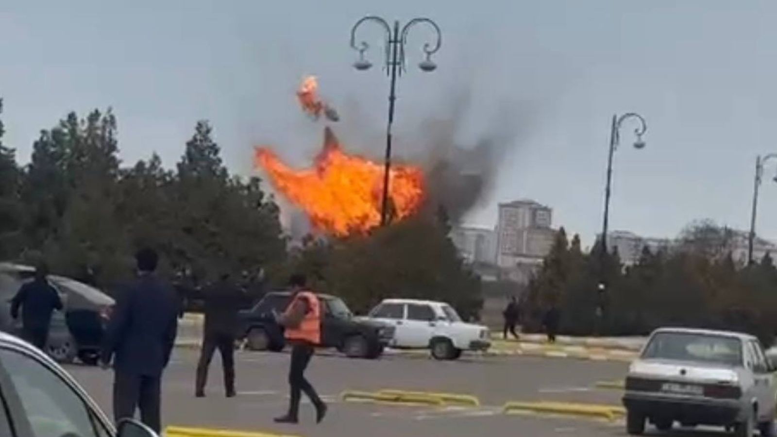 Screenshot of a video showing a large orange fireball from an explosion behind a lamppost in a parking lot. Several people are running away, a person in an orange vest stands near parked Soviet-era cars.