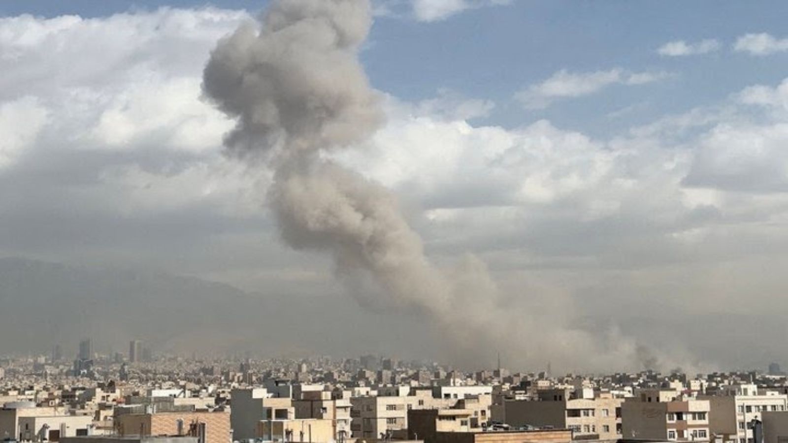 A panoramic view of Tehran with a large plume of gray smoke rising over the city following airstrikes against a cloudy sky.