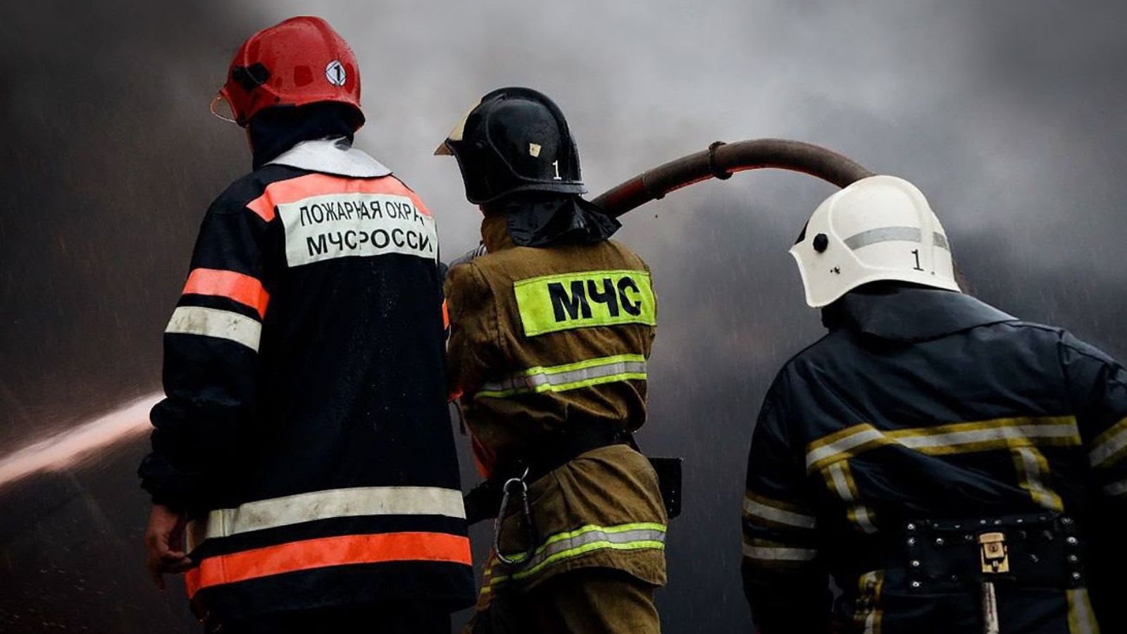 Three firefighters in protective gear extinguishing a large fire against a background of thick smoke.