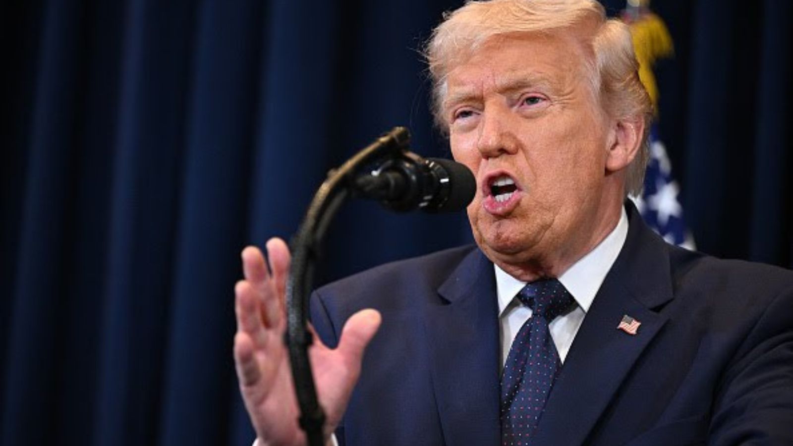 Donald Trump speaking at a podium in front of a blue curtain and the US flag.