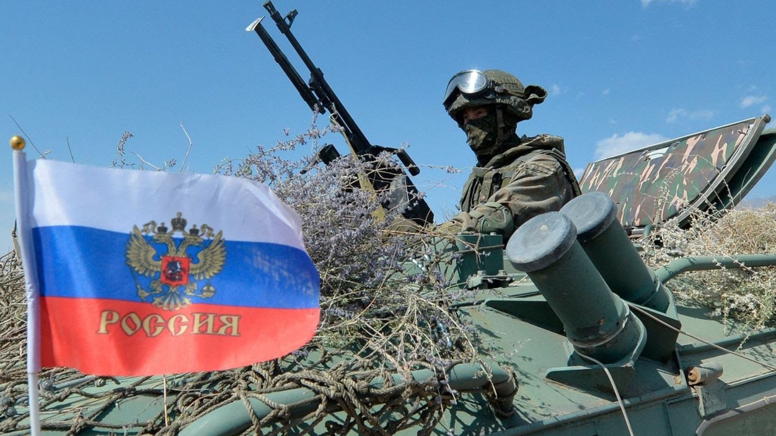A Russian soldier in full combat gear on an armored vehicle next to a Russian flag.