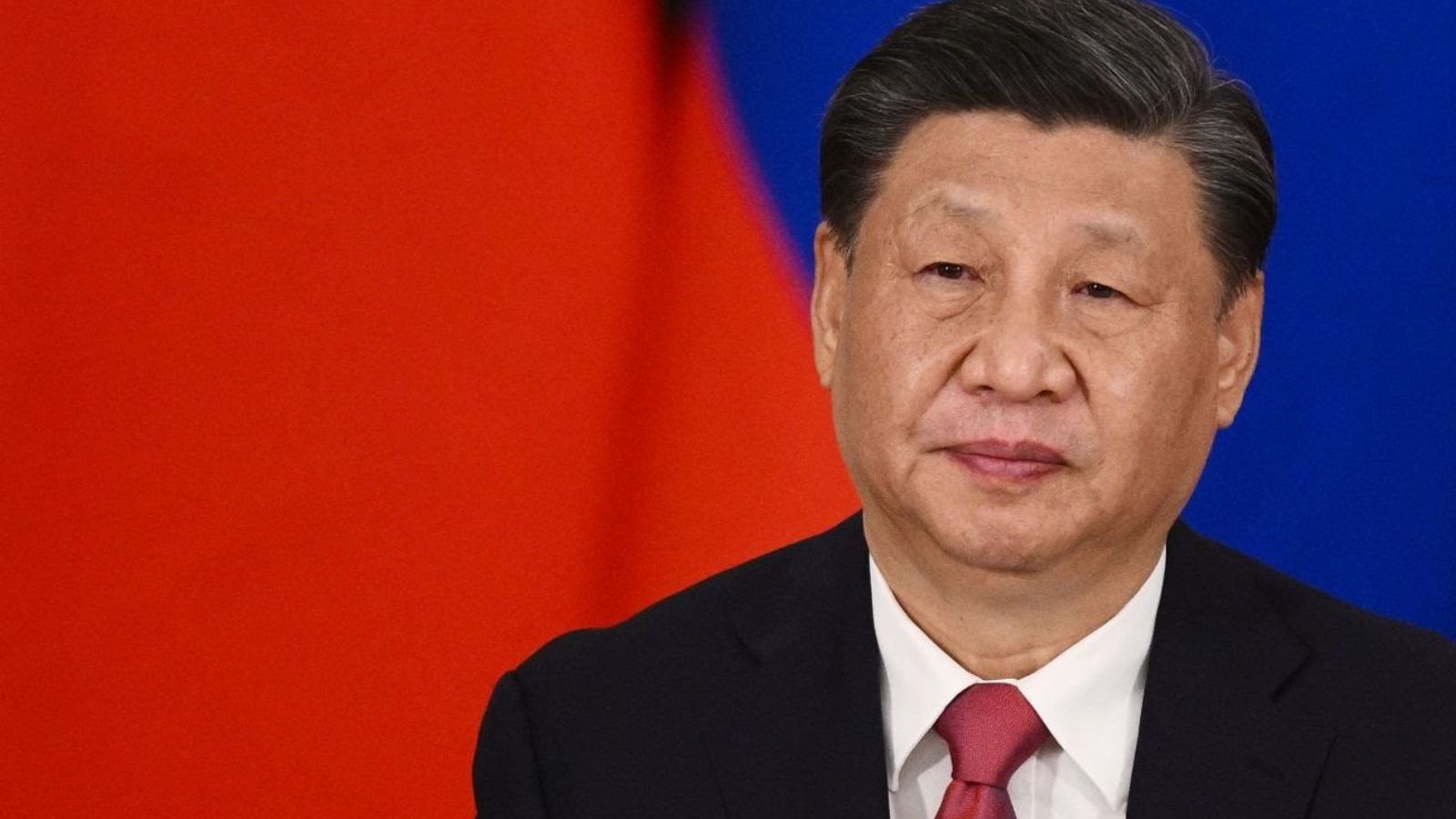 Portrait of Chinese leader Xi Jinping wearing a business suit, positioned against a red and blue background.