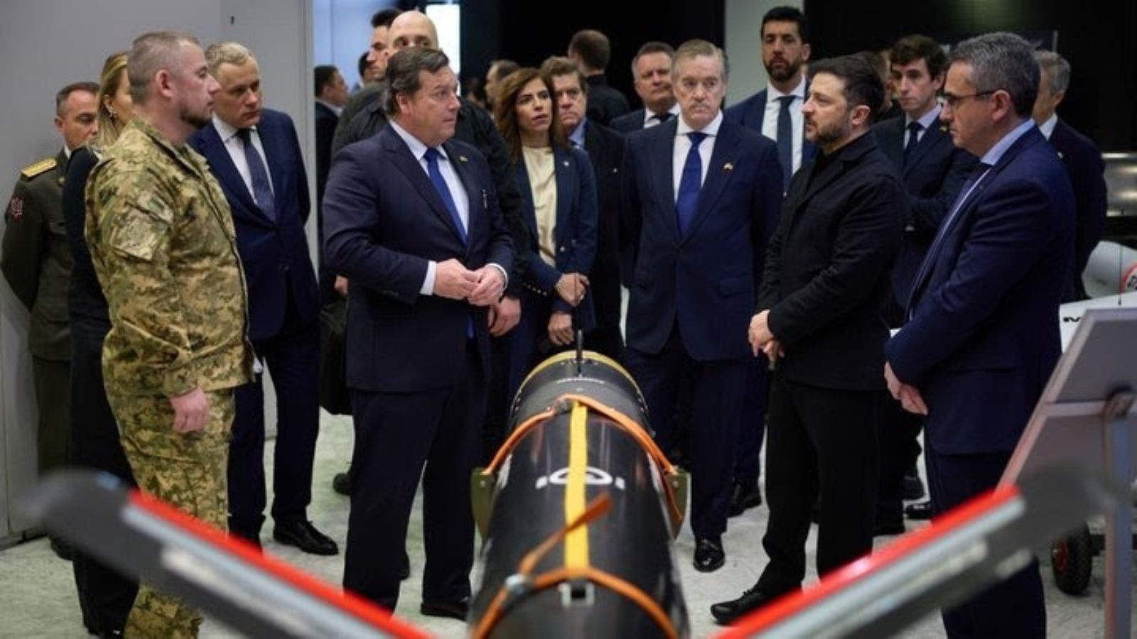 Volodymyr Zelenskyy, dressed in plain black clothing, and a UAF officer in camouflage speak with officials next to large military equipment inside an industrial facility.