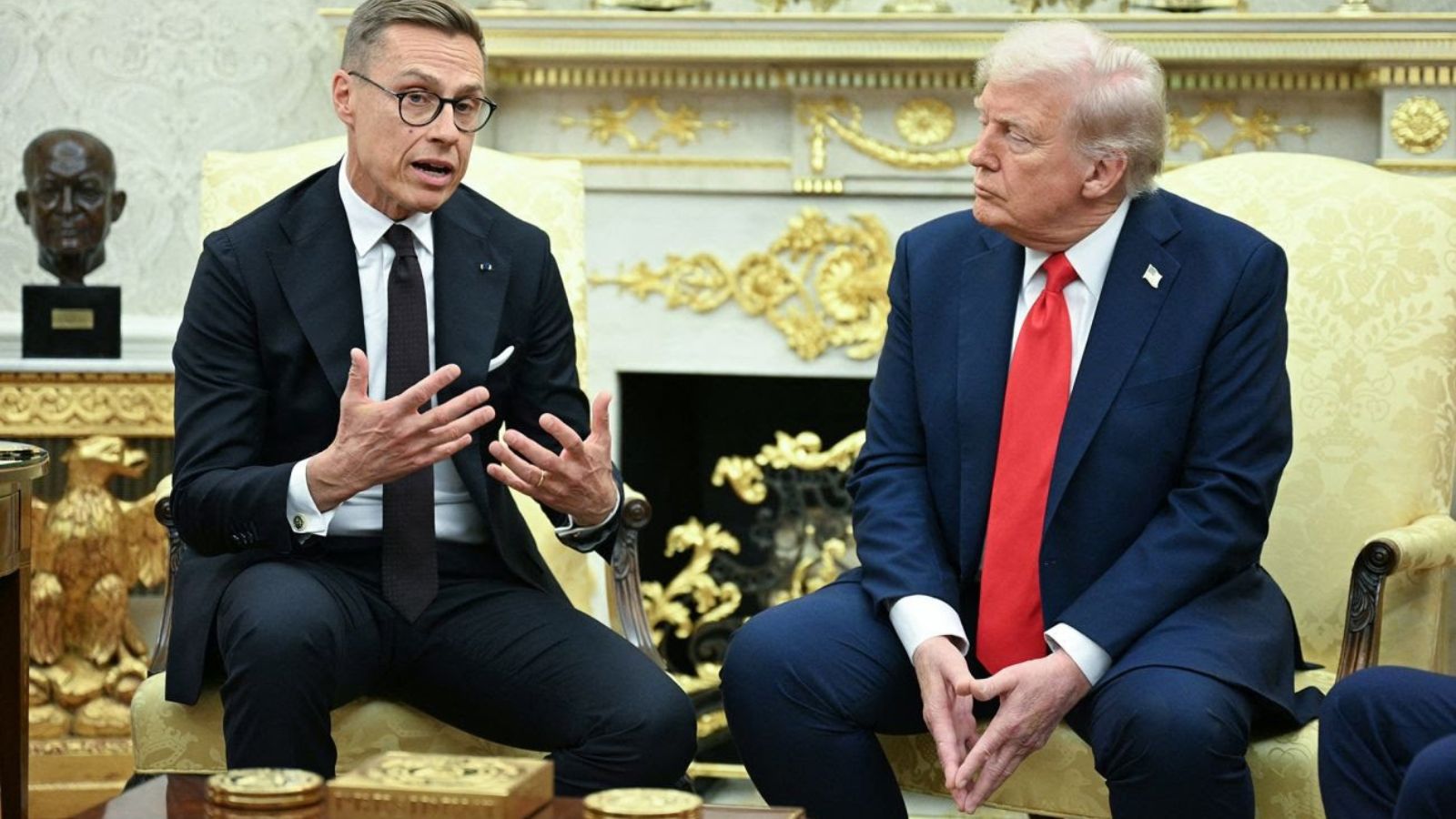 Finnish President Alexander Stubb (left) and US President Donald Trump (right) seated in chairs within the White House during an official meeting. Stubb is speaking with hand gestures, while Trump listens attentively.