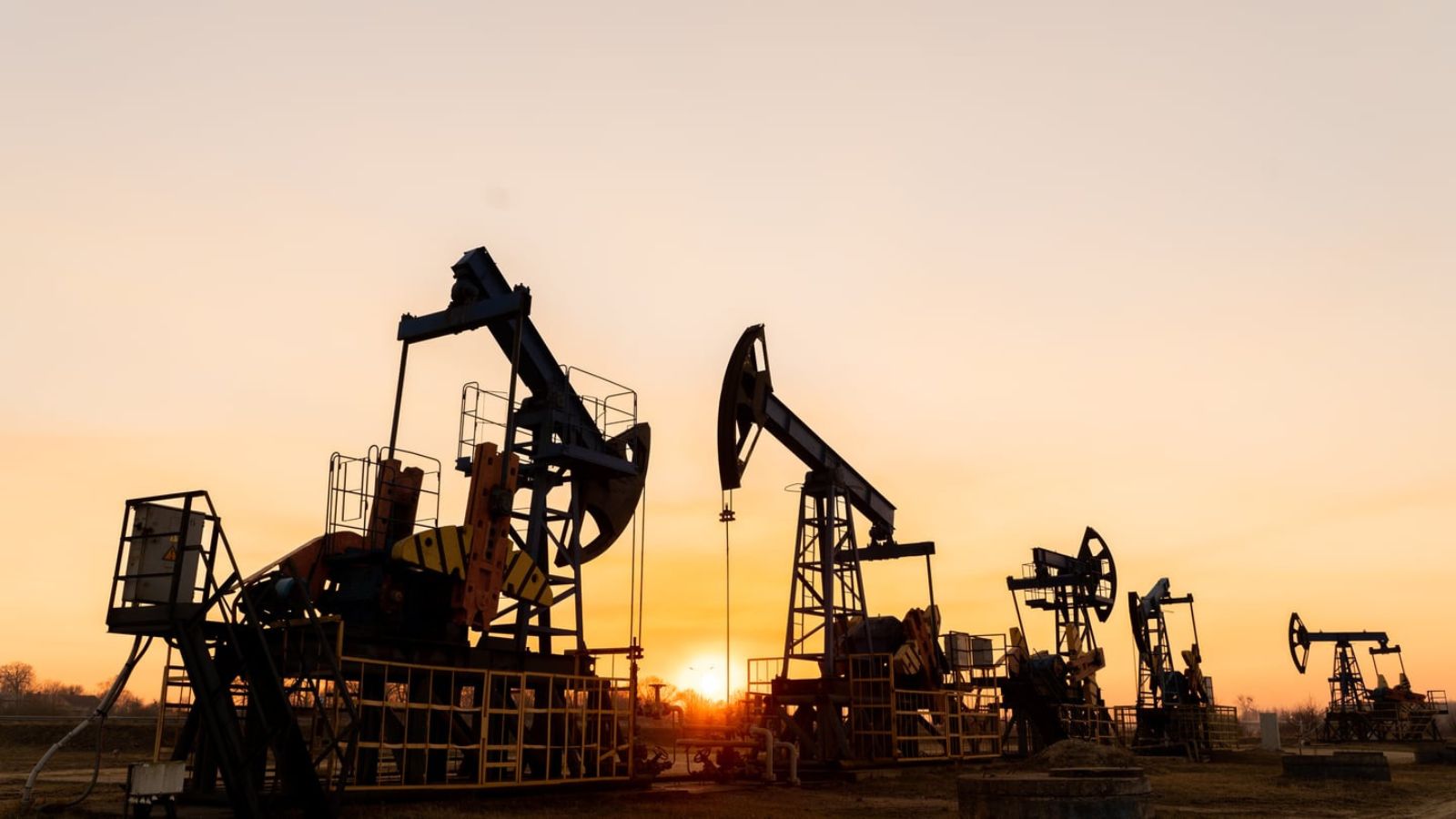 Silhouettes of several working pumpjack oil wells in a field against the setting sun.