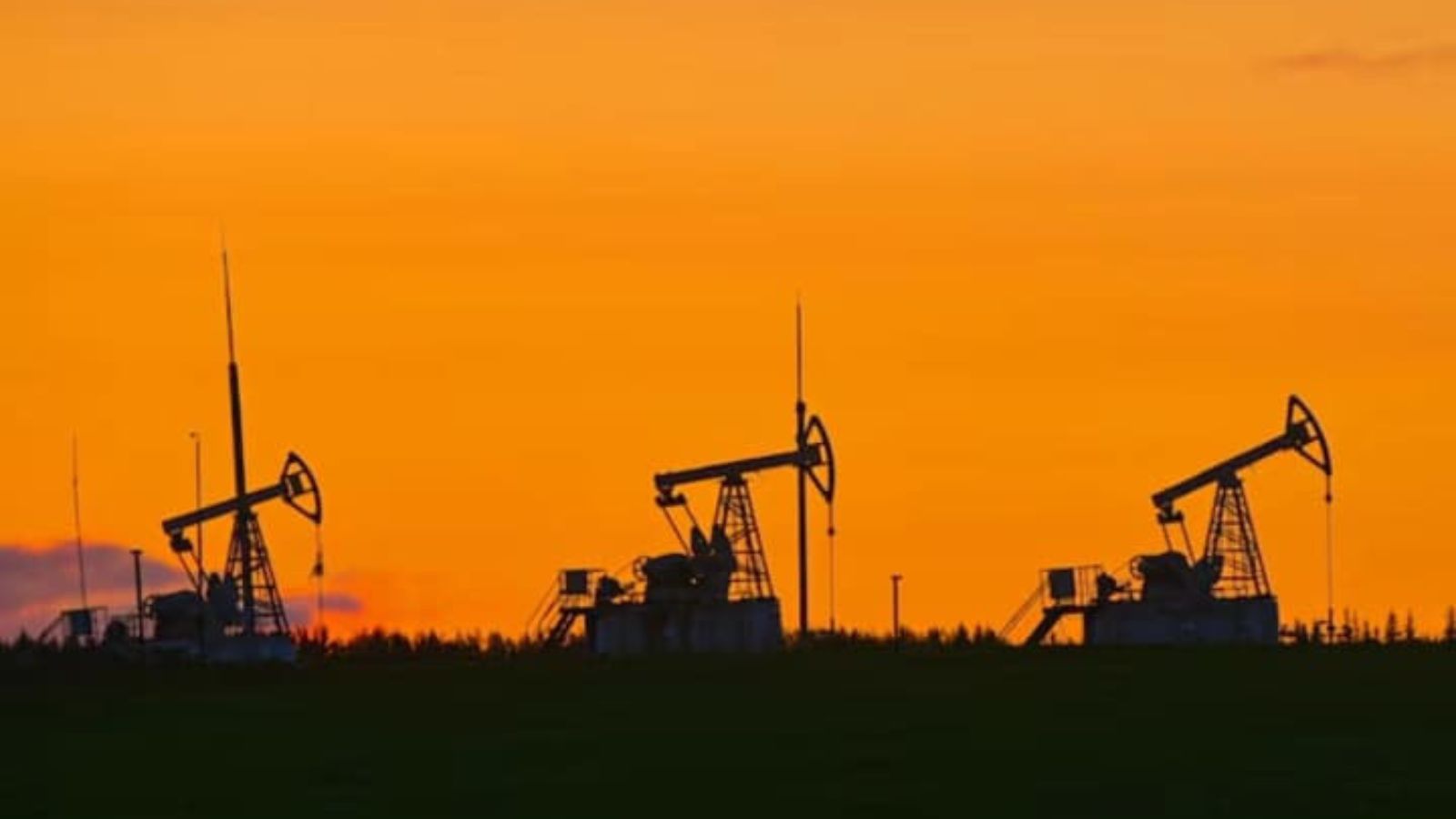 Silhouettes of three operating oil pump jacks on the horizon against a vibrant orange sunset sky