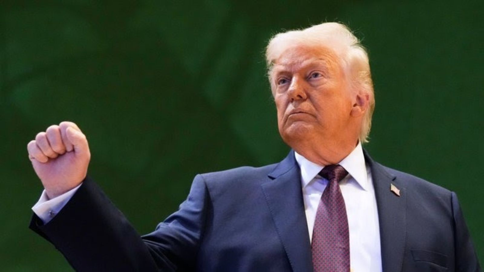 Дональд Трамп у темному костюмі та фіолетовій краватці з рішучим виразом обличчя підняв праву руку в кулаці під час виступу на заході на тлі зеленого фону