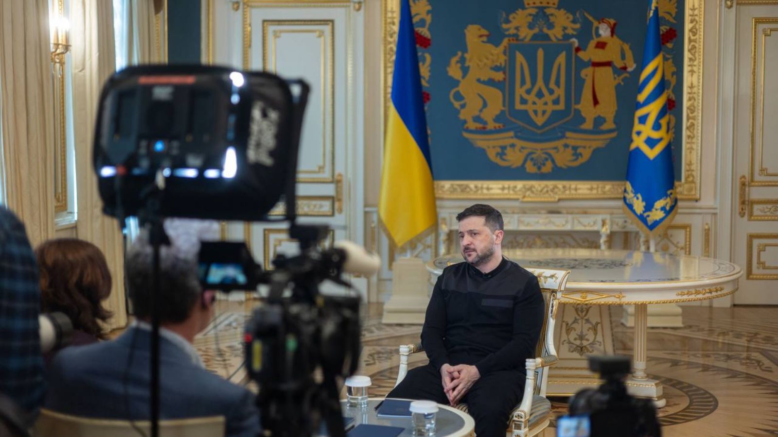 Ukrainian President Volodymyr Zelenskyy is sitting in a gilded armchair at the President’s Office during an interview with Reuters. The Ukrainian flags and coat of arms are behind him. Professional lighting equipment and TV cameras are visible in the foreground.