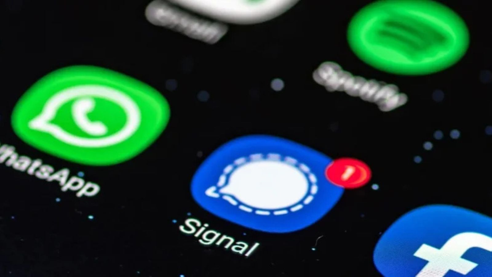 A close-up shot of a smartphone screen displaying the blue Signal app icon with a white chat bubble and a single unread message notification. Part of the WhatsApp and Facebook icons are also visible against a black background.