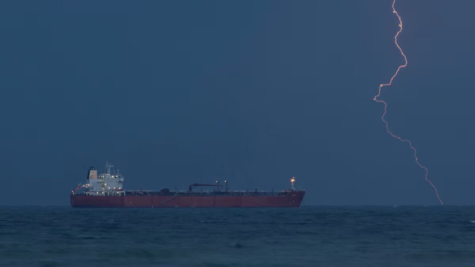 Wide shot: A large red oil tanker sits at anchor in dark waters off the coast of Oman under a stormy sky, with a bright lightning strike hitting in the distance to the right.