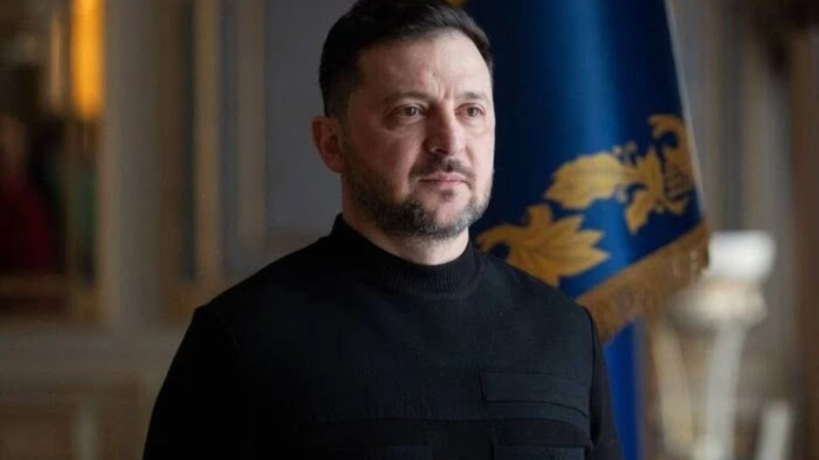 A portrait of the President of Ukraine Volodymyr Zelenskyy. He is wearing a black turtleneck shirt. In the blurred background, the blue and yellow State Flag of Ukraine with golden patterns is visible.