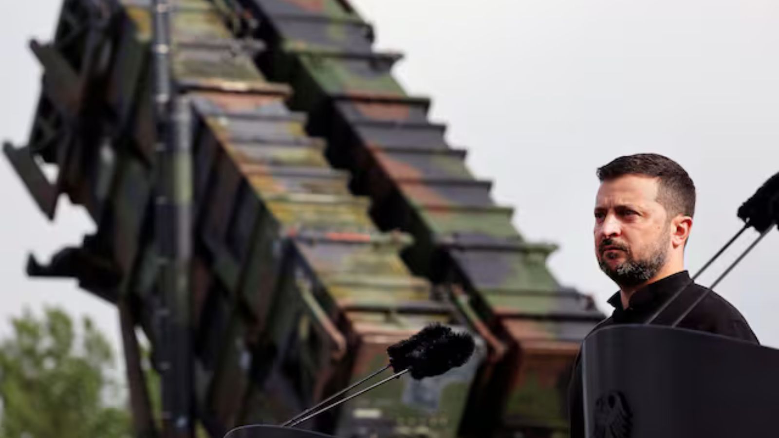 Ukrainian President Volodymyr Zelenskyy, wearing a black shirt, speaks at a podium with microphones, with a massive, camouflaged Patriot missile battery visible in the background.